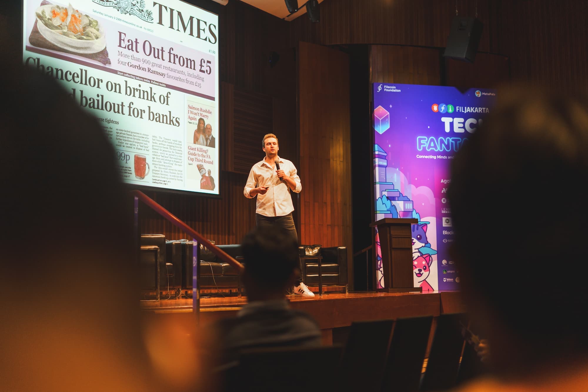 A speaker presenting on stage with a large screen displaying a newspaper front page and a tech event banner.